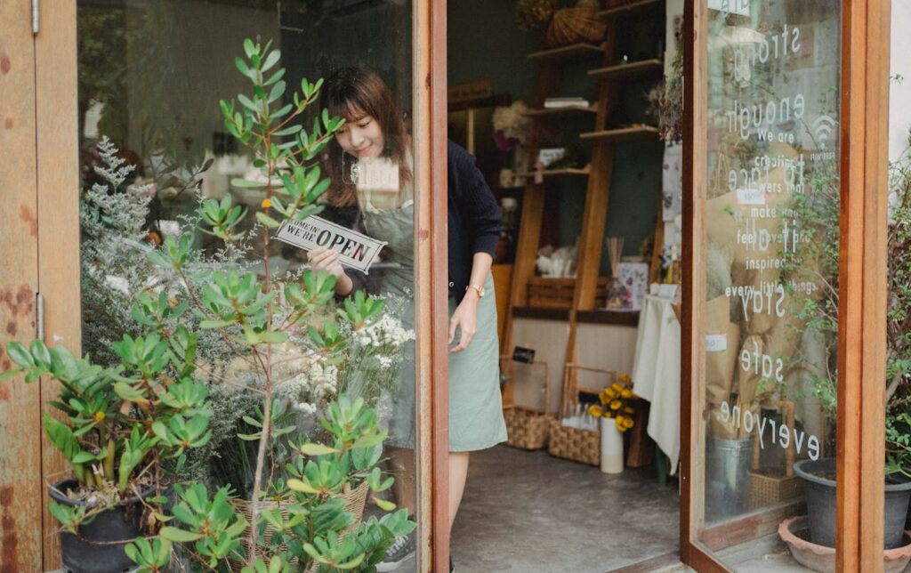 Positive young Asian female florist in apron standing near entrance and turning signboard on glass wall while working in modern floristry shop